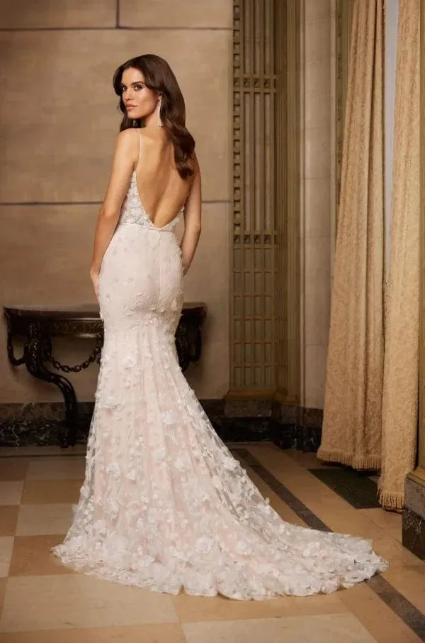 A woman modeling a fitted off-white lace mermaid-style wedding gown with a deep open back, shown from the rear in an elegant indoor room with beige walls and curtains.