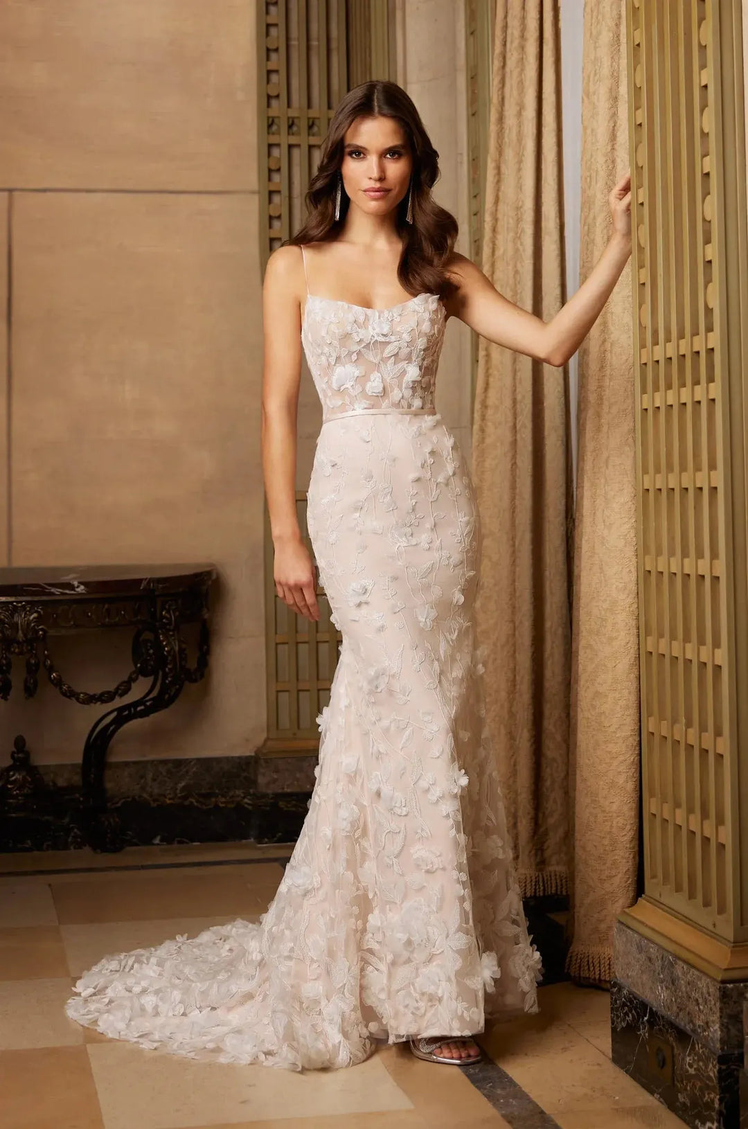 A woman models a strapless mermaid-style wedding gown with a sweetheart neckline, fitted bodice, and cascading floral applique. The dress is a soft champagne or ivory tone with a long train, photographed indoors against beige walls, curtains, and a decorative screen.