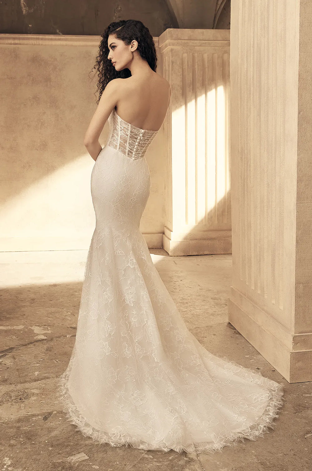 A woman wearing a strapless lace mermaid wedding gown stands in a sunlit architectural interior with stone walls and columns, photographed from the back to show the fitted lace bodice and train.
