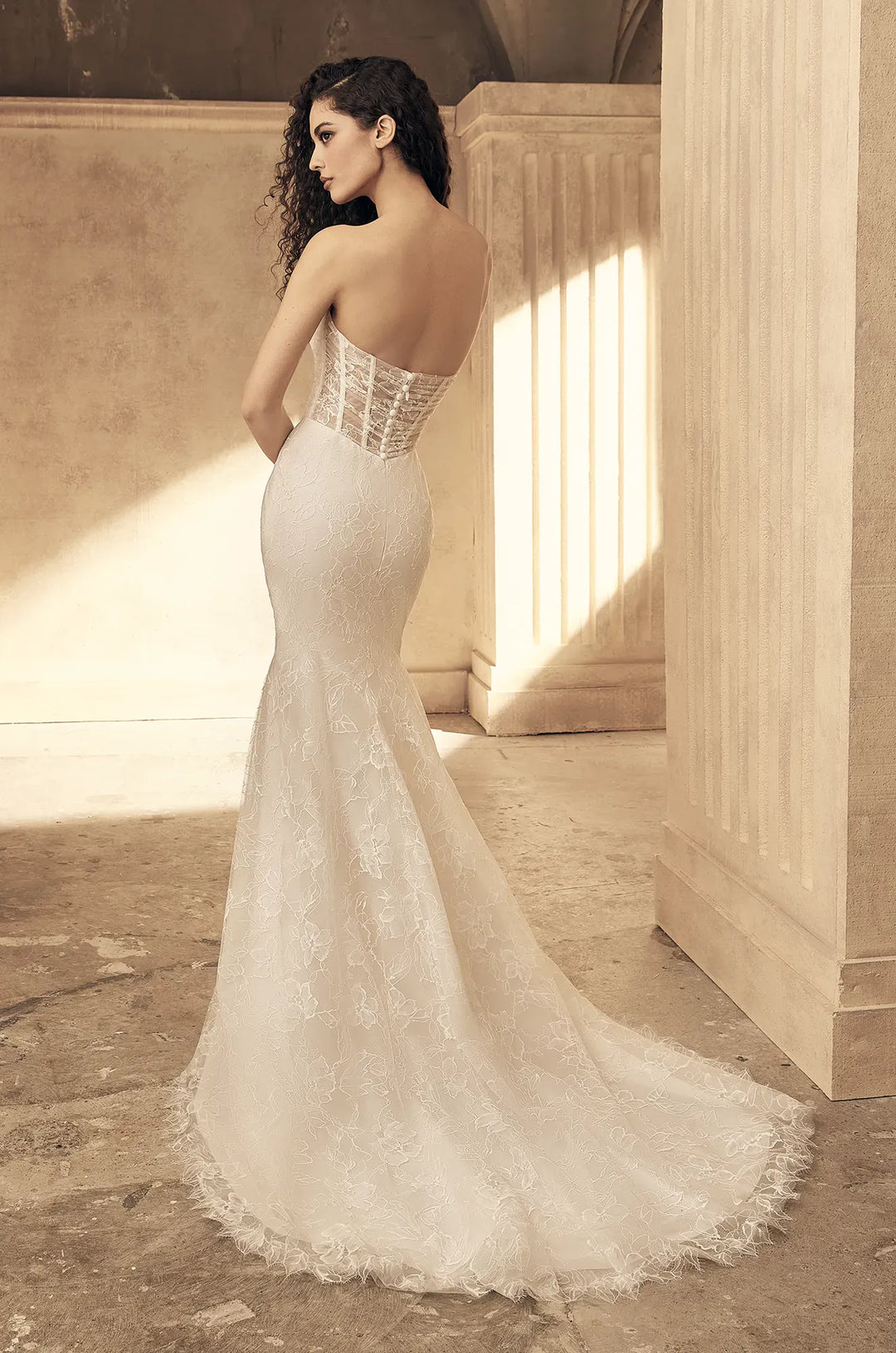 A woman wearing a strapless lace mermaid wedding gown stands in a sunlit architectural interior with stone walls and columns, photographed from the back to show the fitted lace bodice and train.