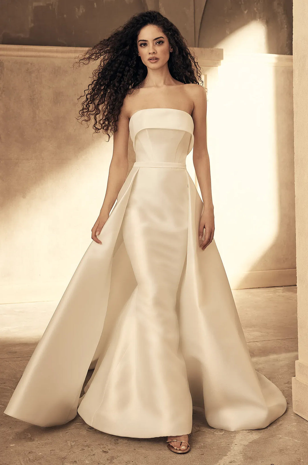 A woman modeling a strapless ivory satin wedding gown with a structured bodice, defined waist, and a full mermaid-style skirt with a train, set against a warm architectural background.