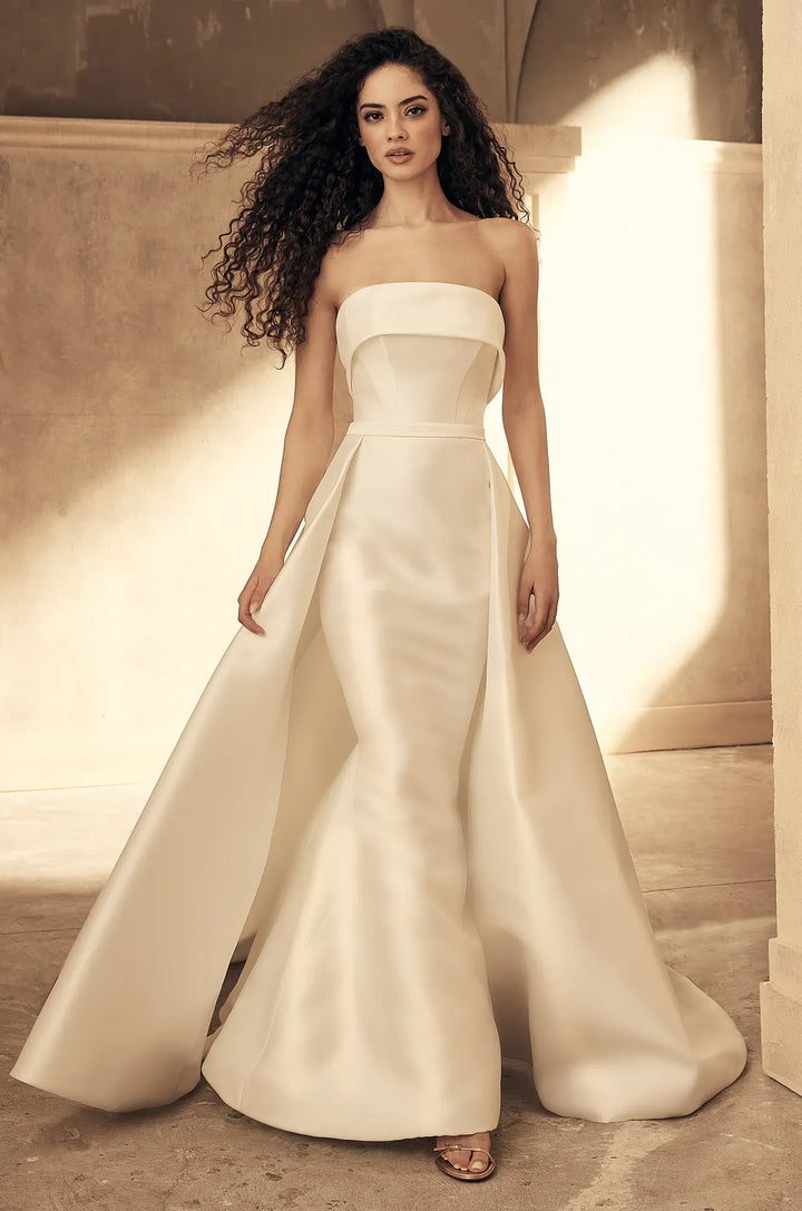 A woman modeling a strapless ivory satin wedding gown with a structured bodice, defined waist, and a full mermaid-style skirt with a train, set against a warm architectural background.