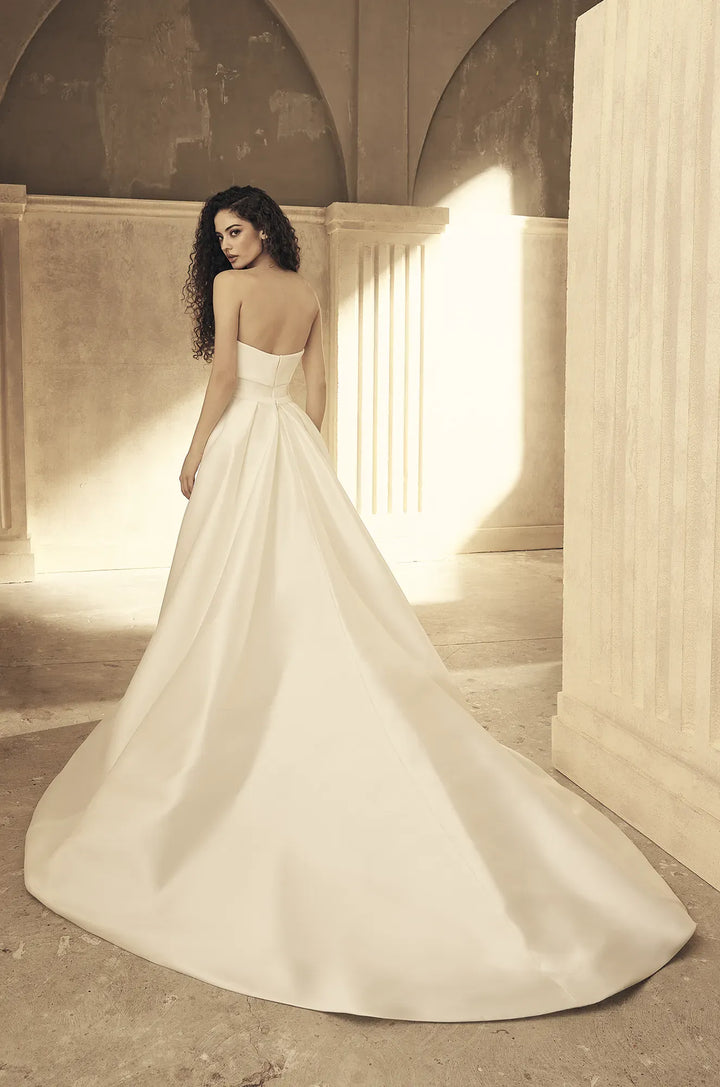 A woman wearing a strapless white wedding gown with a full satin train, photographed in a warm-toned architectural interior with arches and columns.