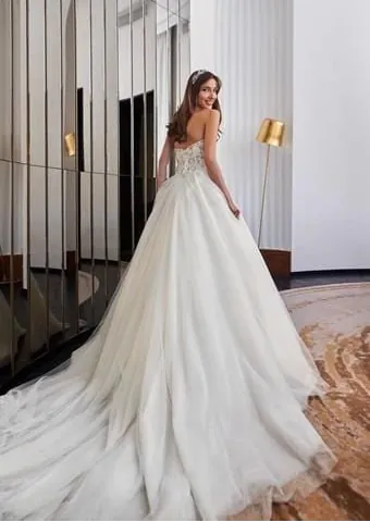 A woman wearing a strapless white wedding gown with a voluminous tulle skirt and long train, standing in a modern interior hallway with mirrored walls and gold floor lamps.