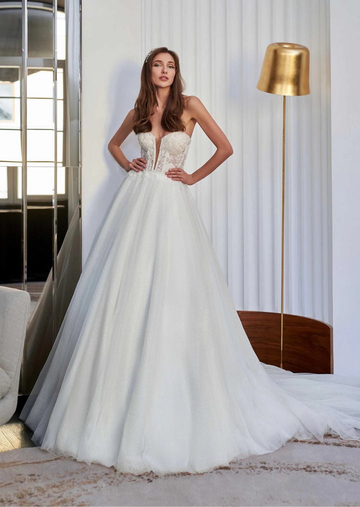 A woman modeling a strapless white ball gown wedding dress with a sheer lace bodice and a full tulle skirt, standing in a bright interior with a gold floor lamp and window visible.
