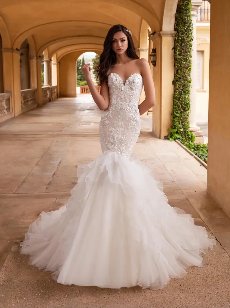 A woman modeling a strapless white mermaid wedding gown with floral lace detailing and a full tulle skirt, standing in a sunlit stone colonnade with arches and ivy.