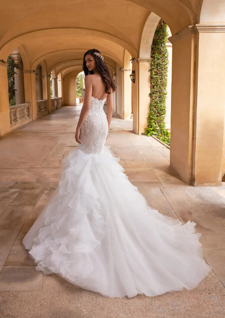 A woman wearing a strapless white mermaid-style wedding gown with a fitted lace bodice and a full layered tulle skirt, standing in a sunlit stone arcade with arches and green ivy.