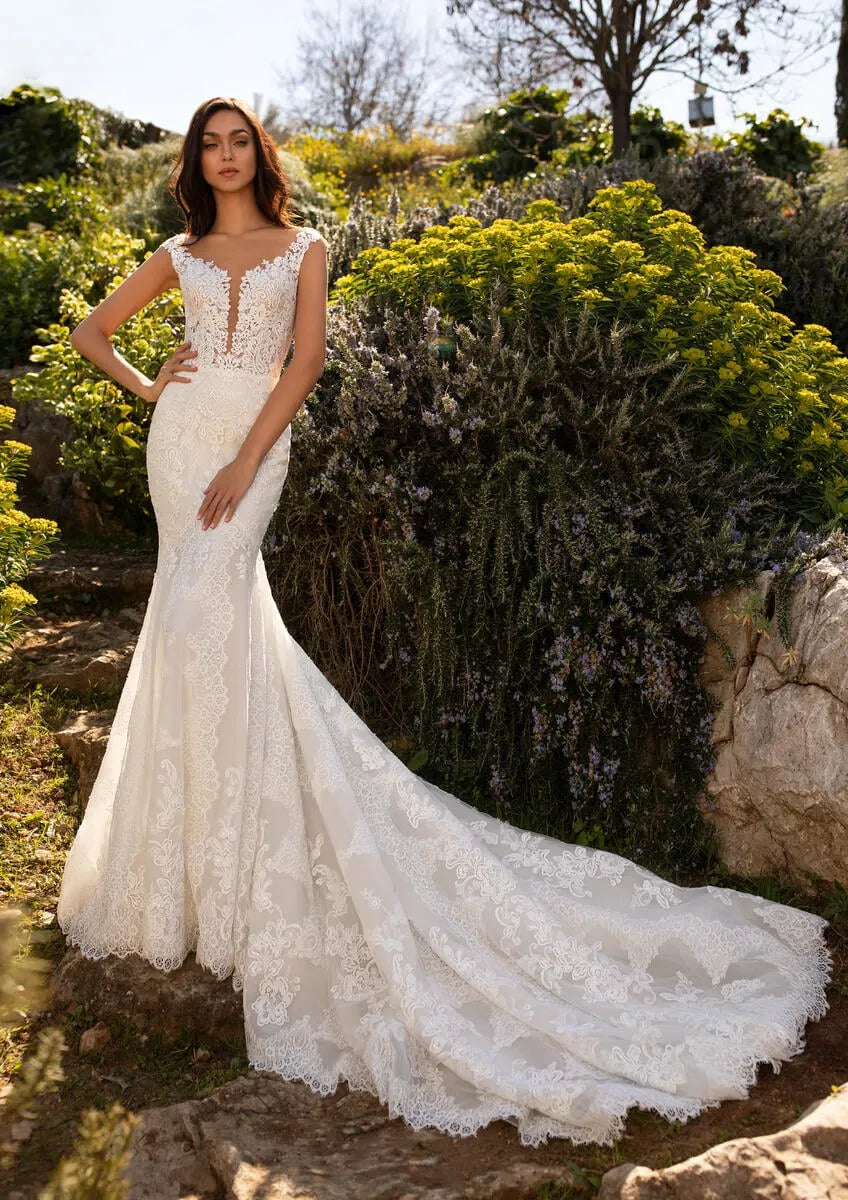 An ivory lace mermaid-style wedding gown with a fitted bodice, illusion neckline, and dramatic train, shown on a model outdoors among rocks and shrubs.