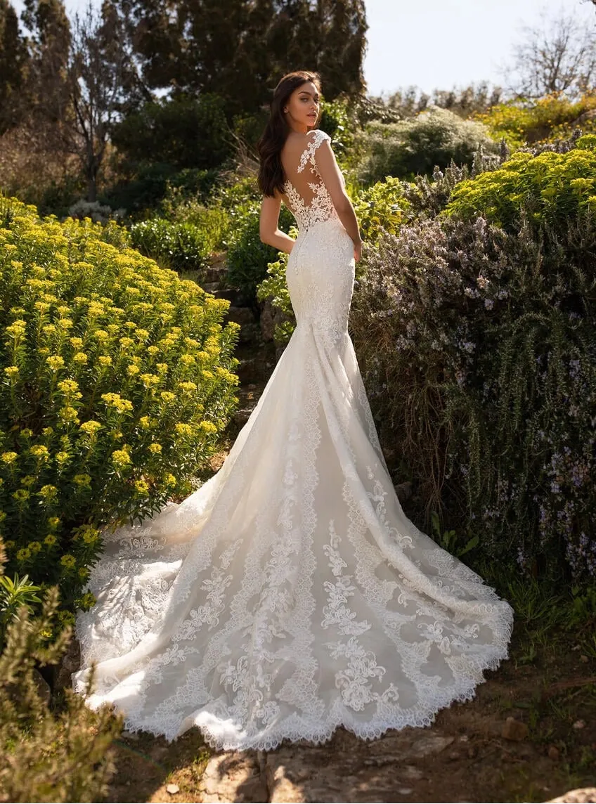 A woman wearing a sleeveless lace mermaid-style wedding gown stands outdoors in a garden, with the full train spread on the ground behind her. The dress has an illusion back and floral lace detailing.