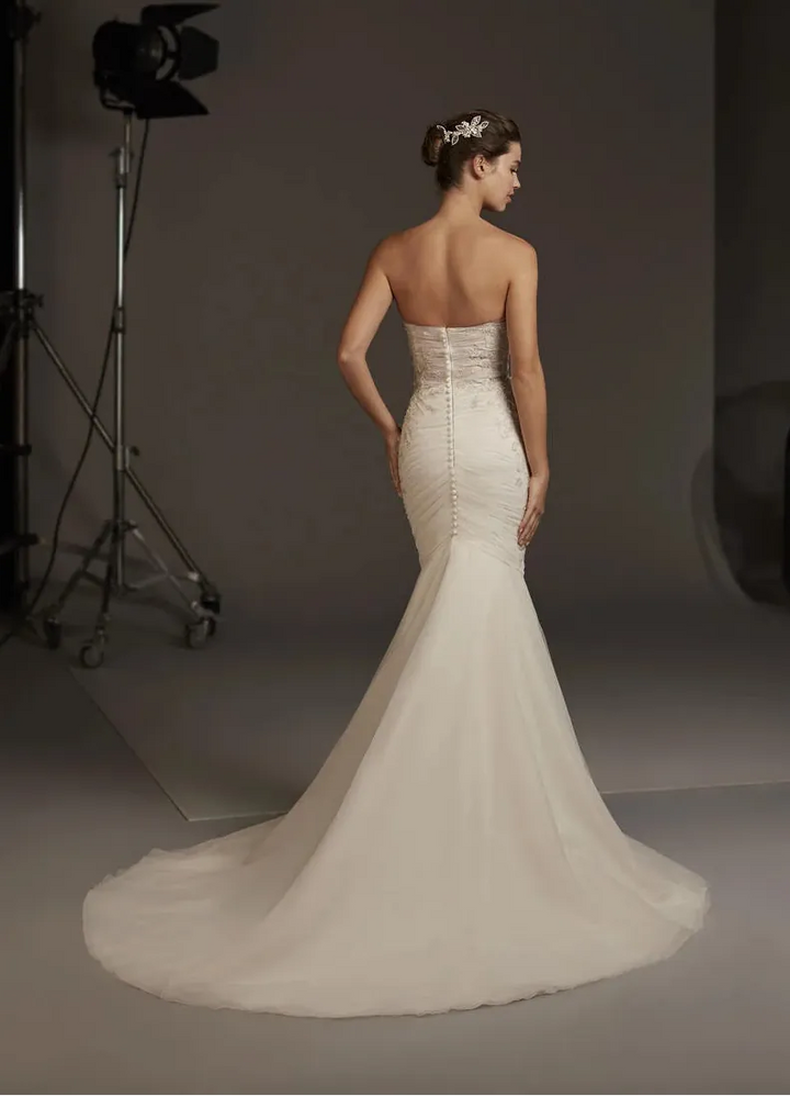 A woman modeling a strapless ivory wedding gown from the back in a studio setting, with a long train and a visible studio light stand in the background.