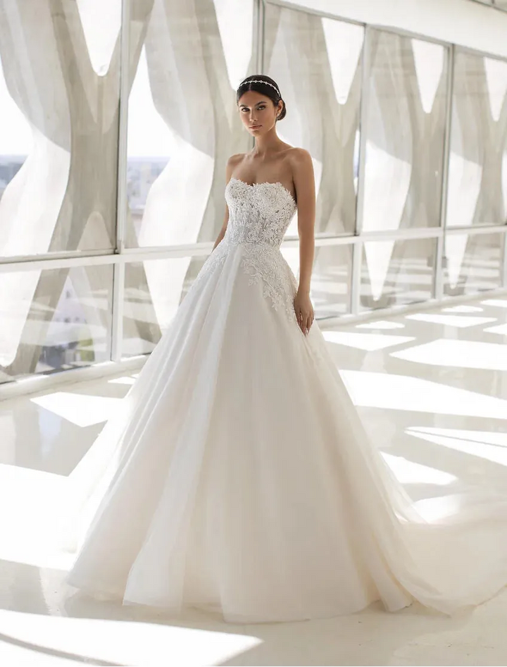 A woman modeling a strapless white wedding gown with a fitted embellished bodice and a full tulle A-line skirt, standing in a bright modern interior with large windows and sheer drapes.