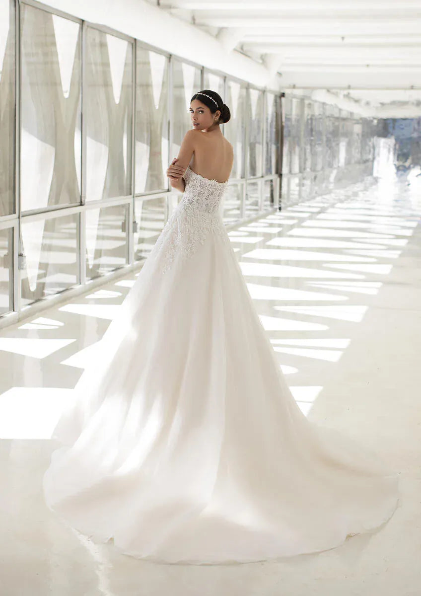 A woman wearing a strapless white wedding gown with a fitted lace bodice and a full flowing skirt, standing in a bright modern corridor with large windows.