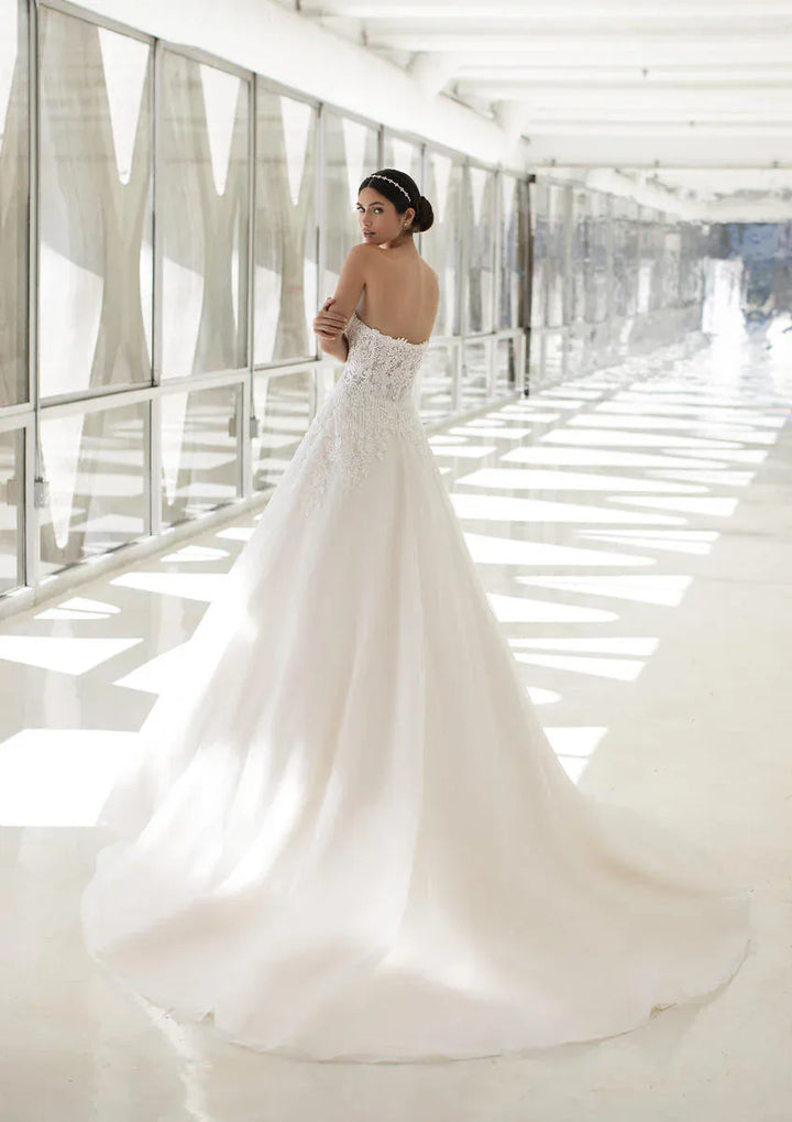 A woman wearing a strapless white wedding gown with a fitted lace bodice and a full flowing skirt, standing in a bright modern corridor with large windows.