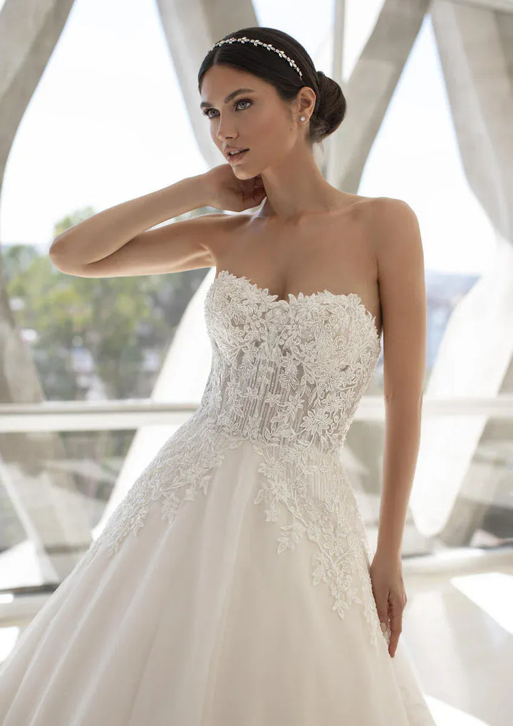 A woman modeling a strapless white wedding gown with a sweetheart neckline, heavily embellished lace and beadwork on the bodice, and a full skirt, posed in front of a bright, modern architectural background.