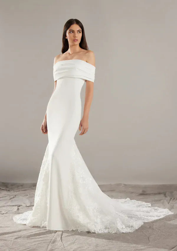 An off-the-shoulder white wedding gown with a fitted silhouette and a lace-trimmed mermaid-style hem, shown on a model against a neutral studio backdrop.