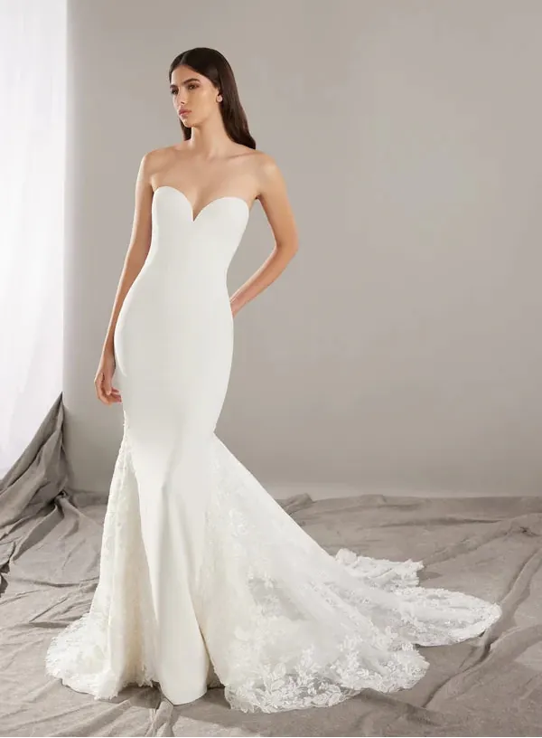 A woman modeling a strapless white mermaid-style wedding gown with a sweetheart neckline, fitted bodice, lace flared hem, and long train in a softly lit studio with a neutral gray backdrop.