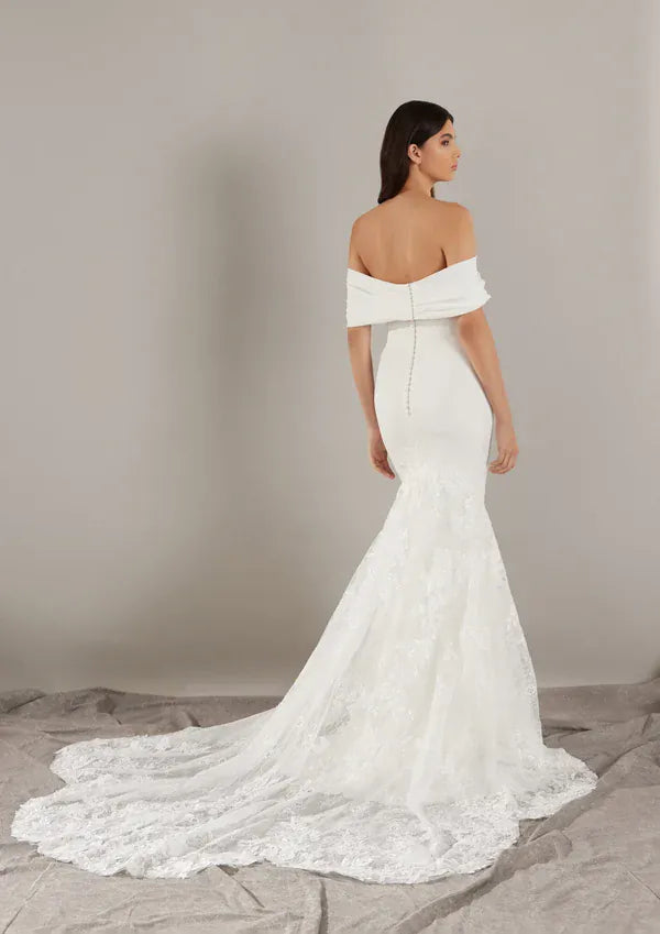 A woman modeling an off-the-shoulder white lace mermaid wedding dress with a long train, photographed from the back against a neutral gray studio backdrop.