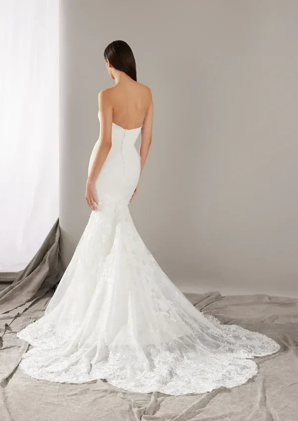 A woman modeling a strapless white mermaid-style wedding dress from the back, standing on a draped gray fabric floor against a light gray studio wall.