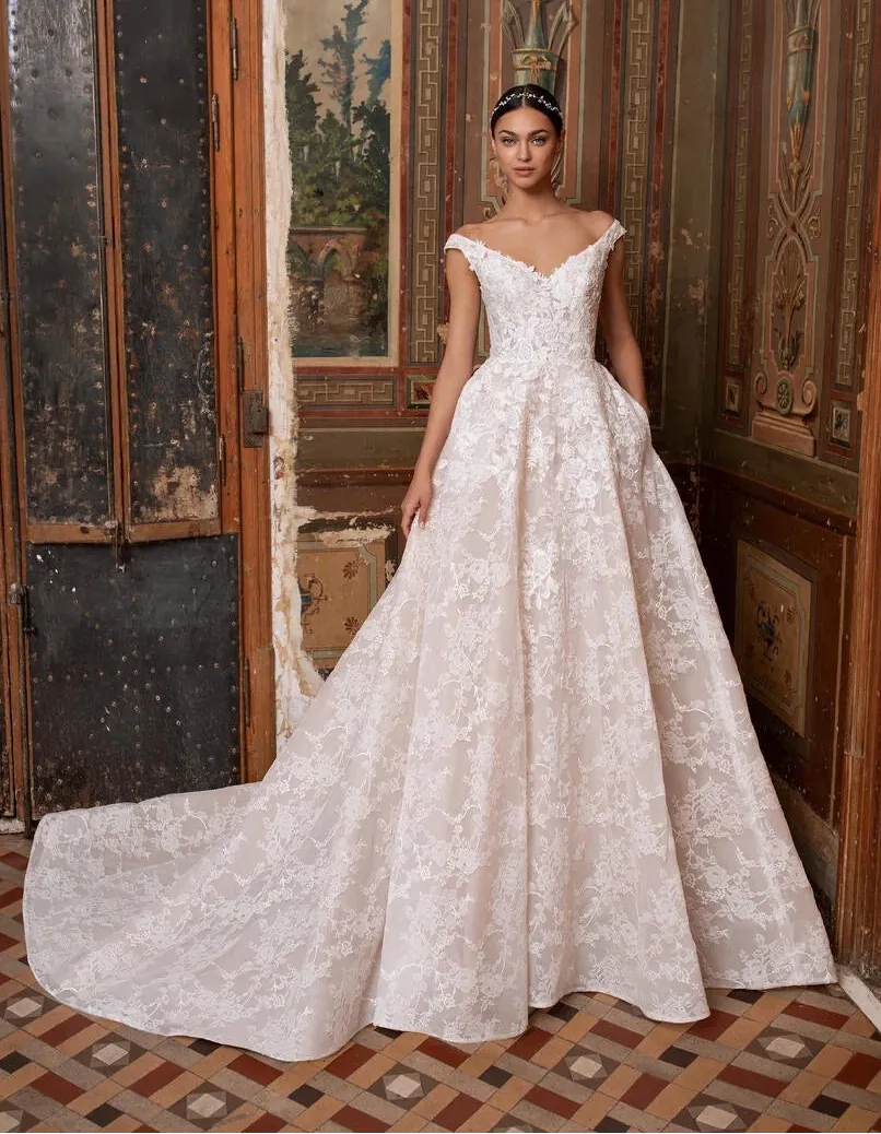 A woman wearing a voluminous off-white lace ball gown with a train stands in an ornate interior with carved wood doors and tiled flooring.