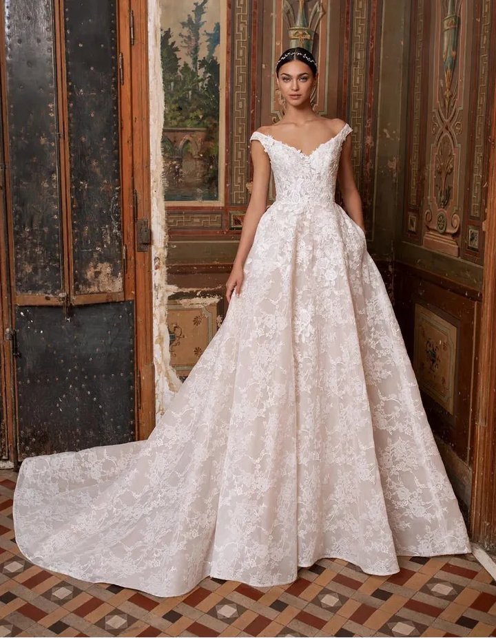 A woman wearing a voluminous off-white lace ball gown with a train stands in an ornate interior with carved wood doors and tiled flooring.