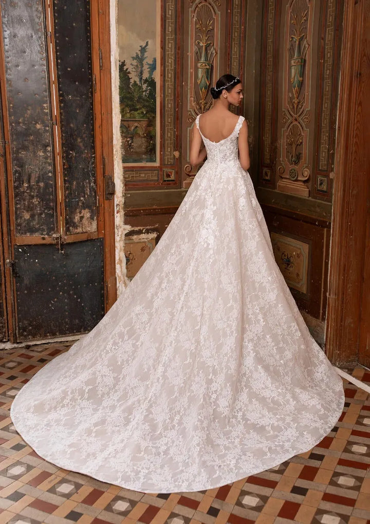 A woman wearing a voluminous white lace wedding gown with a long train, photographed from behind in an ornate interior with wooden doors and patterned tile flooring.