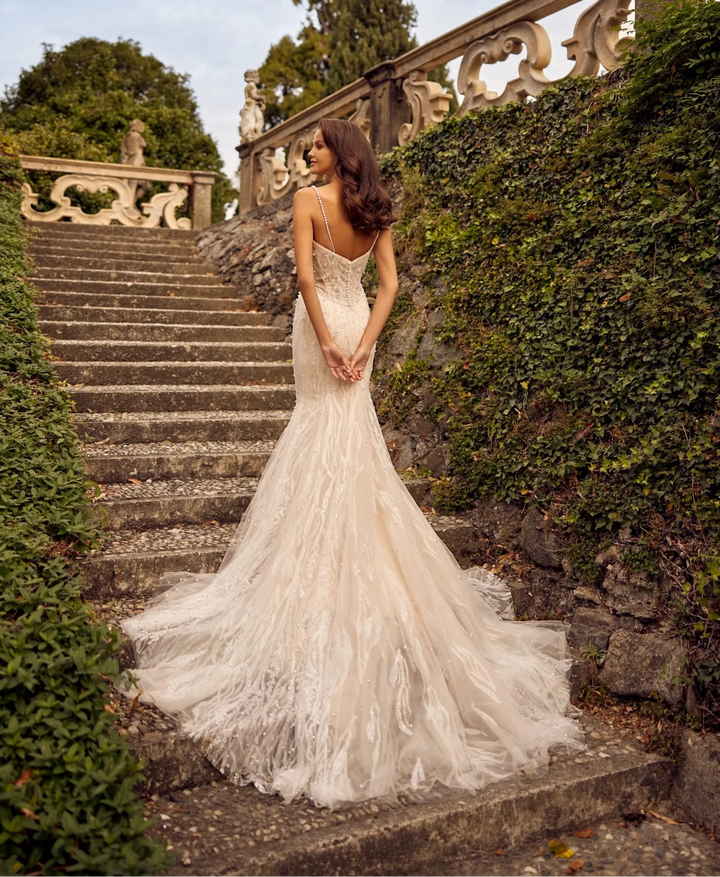 A woman wearing a strapless ivory lace mermaid wedding gown stands on stone stairs between green ivy-covered walls, photographed from behind as the dress train spreads across the steps.