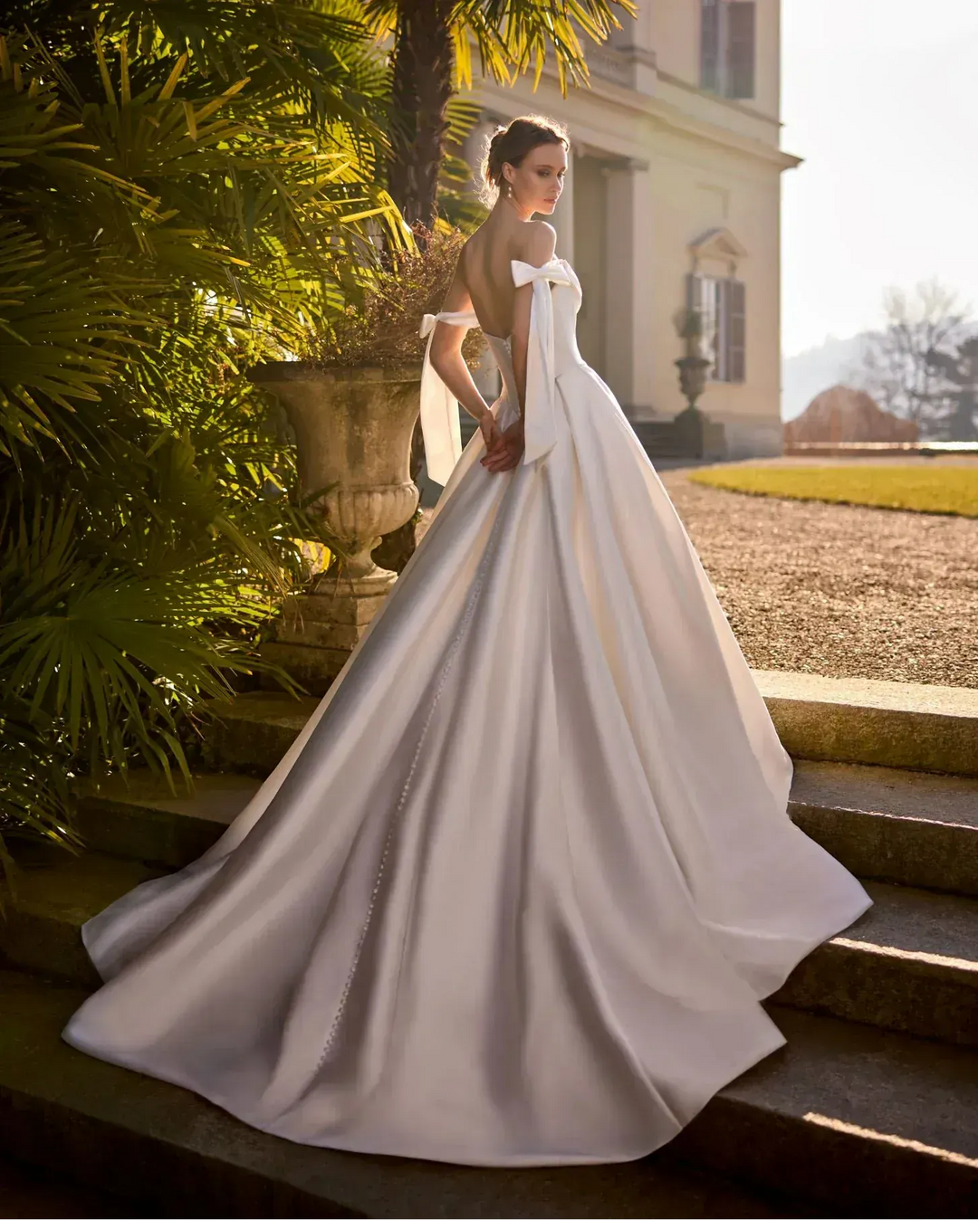 Woman in a white wedding dress standing on steps with a classical building and garden in the background