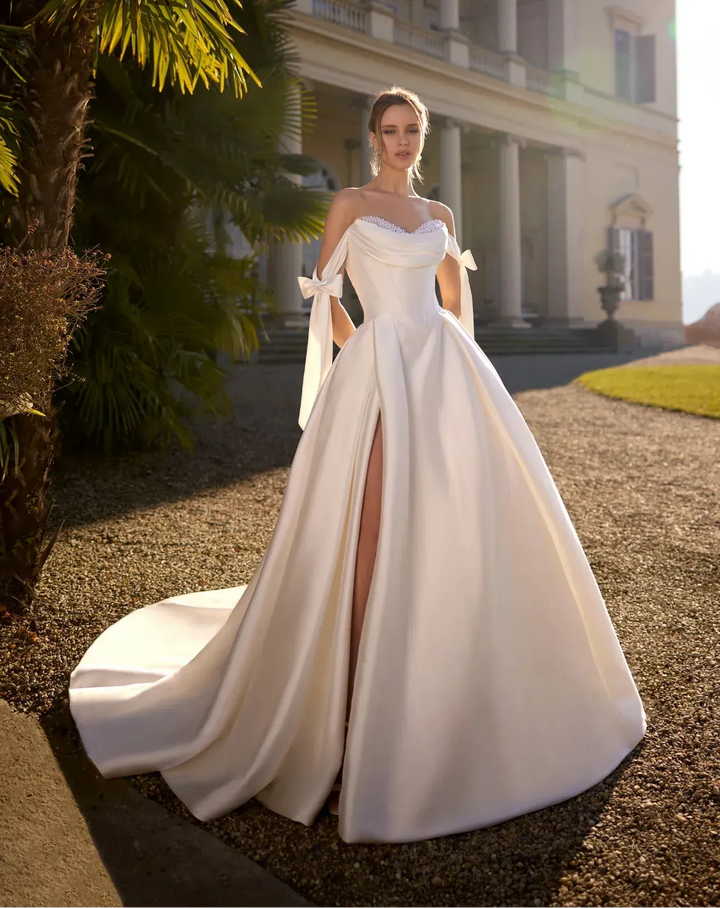 A woman wearing a white off-the-shoulder ball gown with a high thigh slit stands outdoors in front of a grand classical building, with palm trees and warm sunlight on the scene.