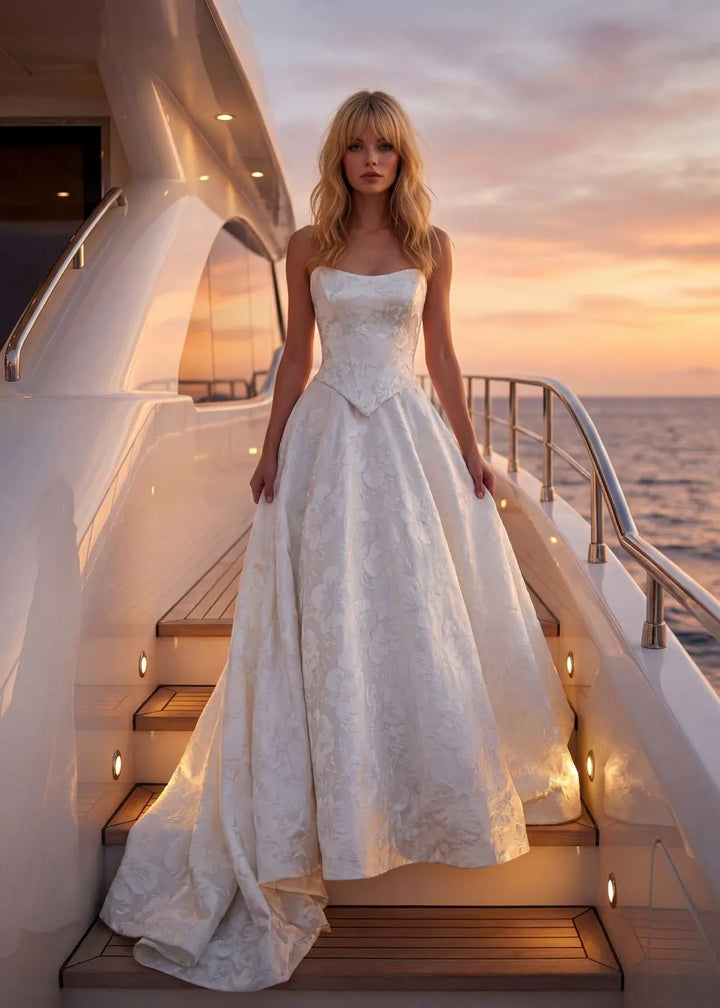 A woman wearing a strapless white wedding gown with a fitted bodice and full skirt stands on the stairs of a yacht at sunset, with the ocean and sky visible in the background.
