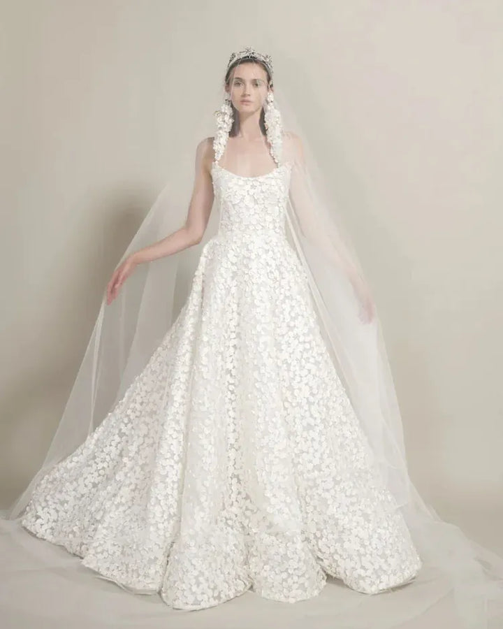 A model wearing a voluminous white floral lace wedding gown with a sheer veil and floral headpiece, posed against a neutral beige studio backdrop.