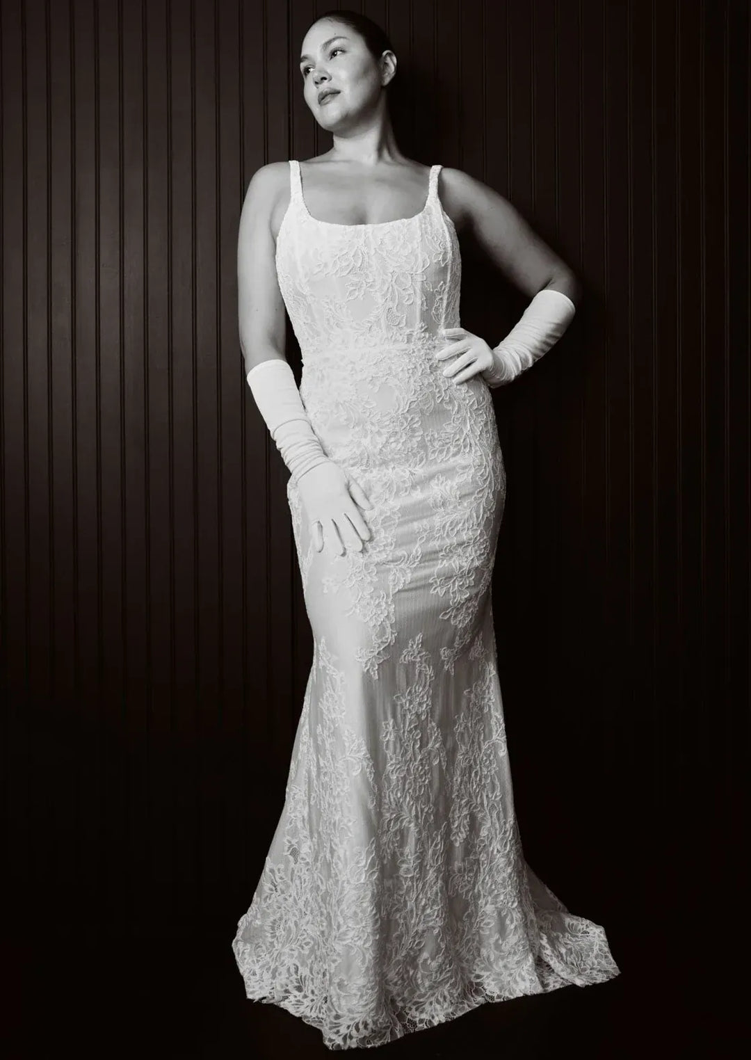 A woman wearing a fitted white lace evening gown with thin straps, long white opera gloves, and a slight train, posed against a dark vertically paneled background.