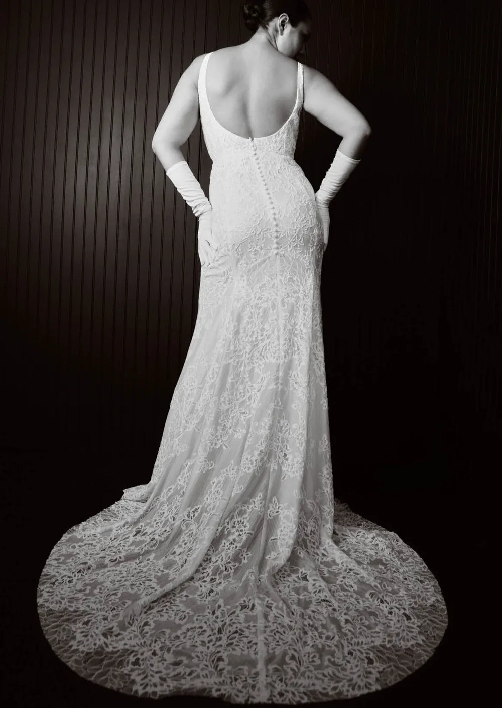 A woman photographed from behind wearing a fitted lace wedding gown with a deep open back, long gloves, and a long train against a dark paneled background.