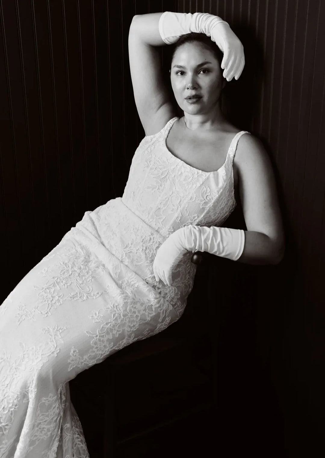 A black and white fashion photograph of a woman wearing a sleeveless lace wedding gown and matching long gloves, leaning against a dark paneled wall.