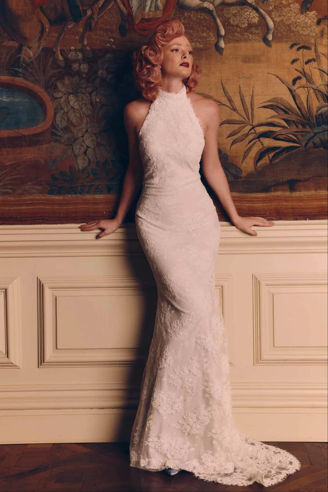 A woman wearing a fitted white lace halter gown with a long train, posed against an ornate wall with a large painted mural in the background.