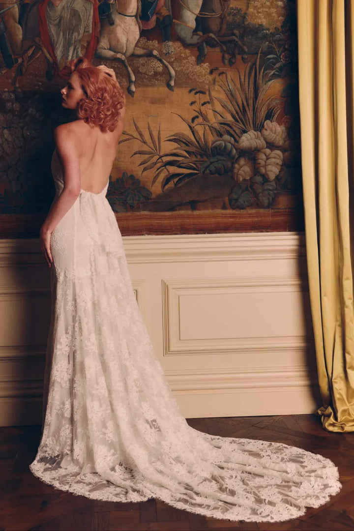 A woman wearing a white lace wedding gown with a long train, photographed indoors against a large tapestry and gold curtain.