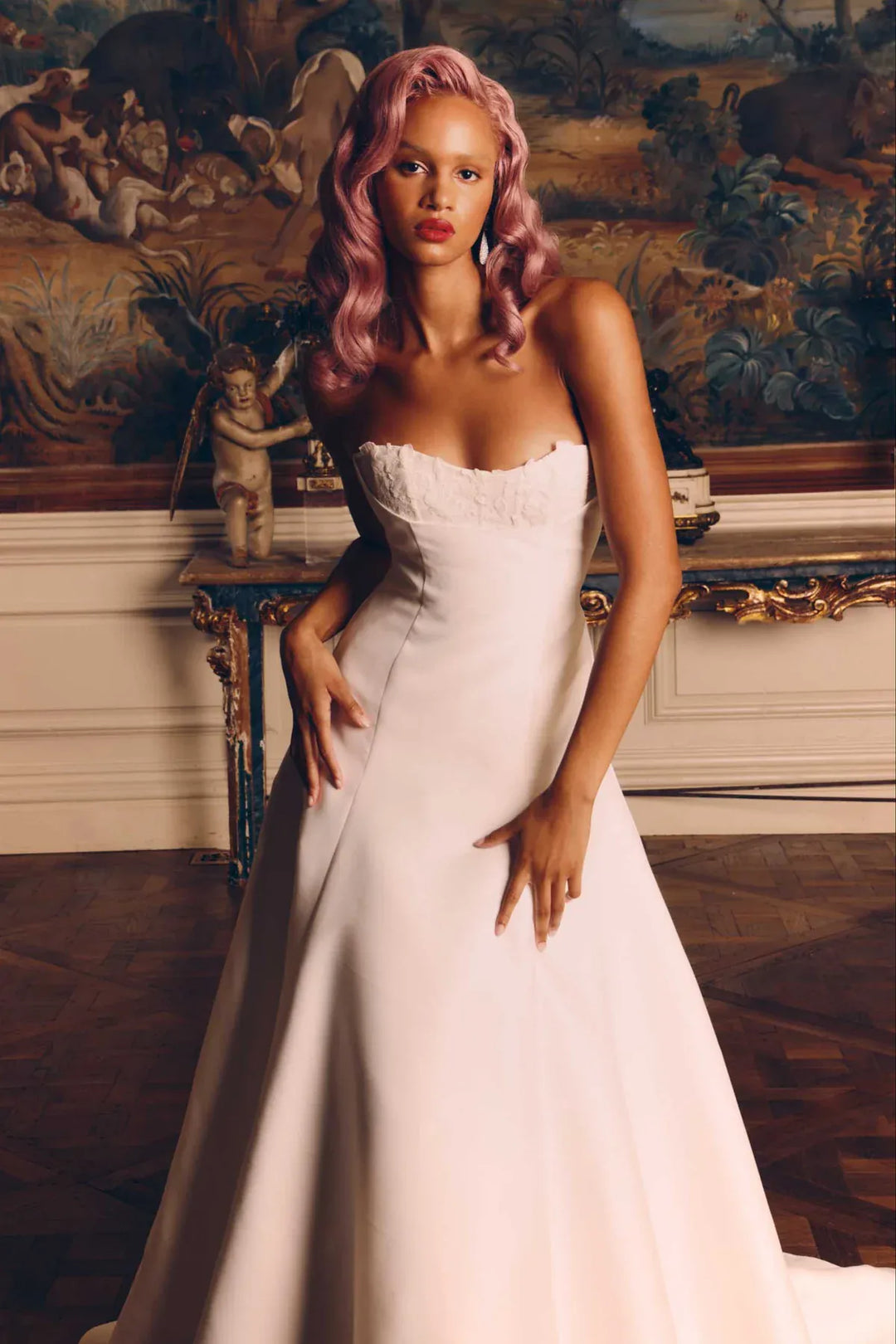 Woman in a white strapless gown with pink hair standing in an ornate room.