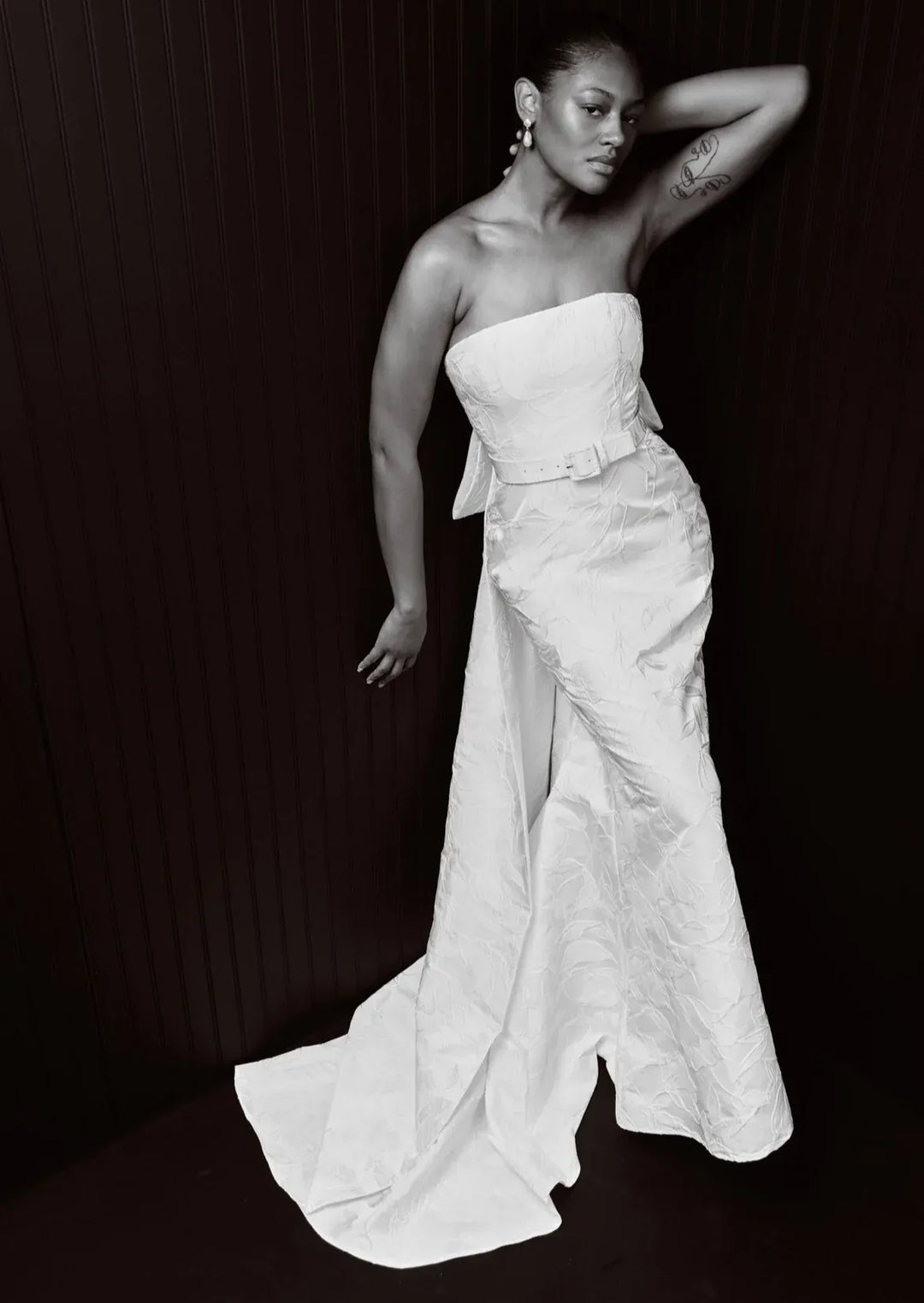 A black and white fashion photograph of a model wearing a strapless white gown with a structured bodice, waist bow, and a long flowing train against a dark paneled background.