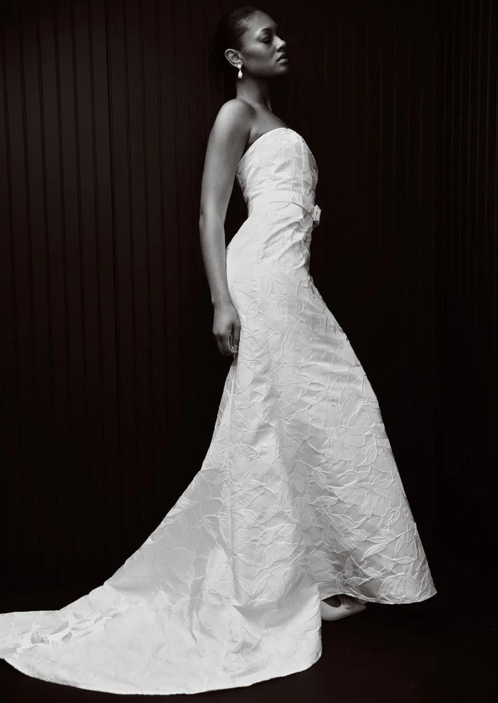 A woman in profile wearing a strapless white textured gown with a fitted bodice and a long train, posed against a dark paneled background.