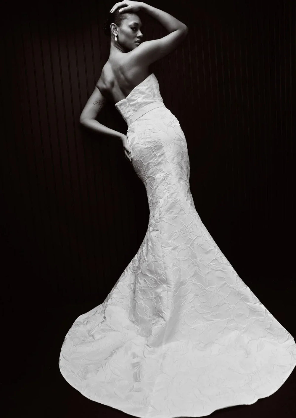 A black and white fashion photograph of a model wearing a strapless mermaid-style wedding gown with a textured, crinkled fabric and a full train against a dark background.