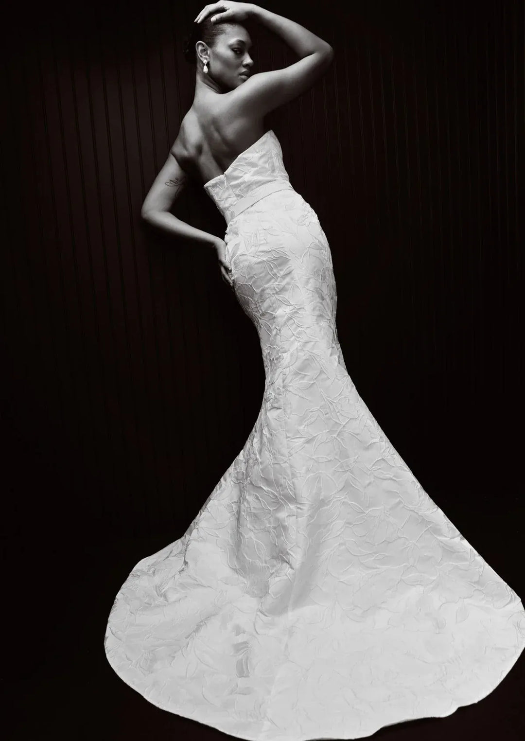 A black and white fashion photograph of a model wearing a strapless mermaid-style wedding gown with a textured, crinkled fabric and a full train against a dark background.