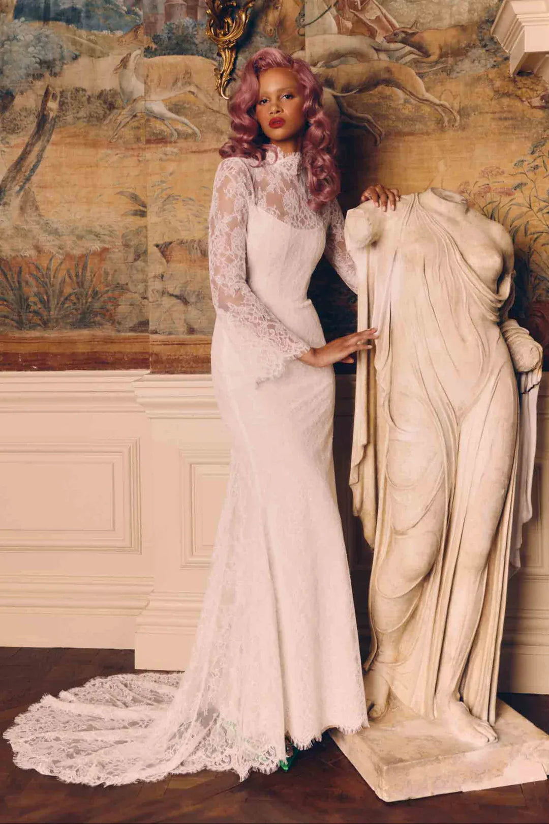 A woman with pink hair wears a fitted white lace mermaid gown with long sheer sleeves and a high neckline, standing indoors beside a large classical statue against a painted mural wall.