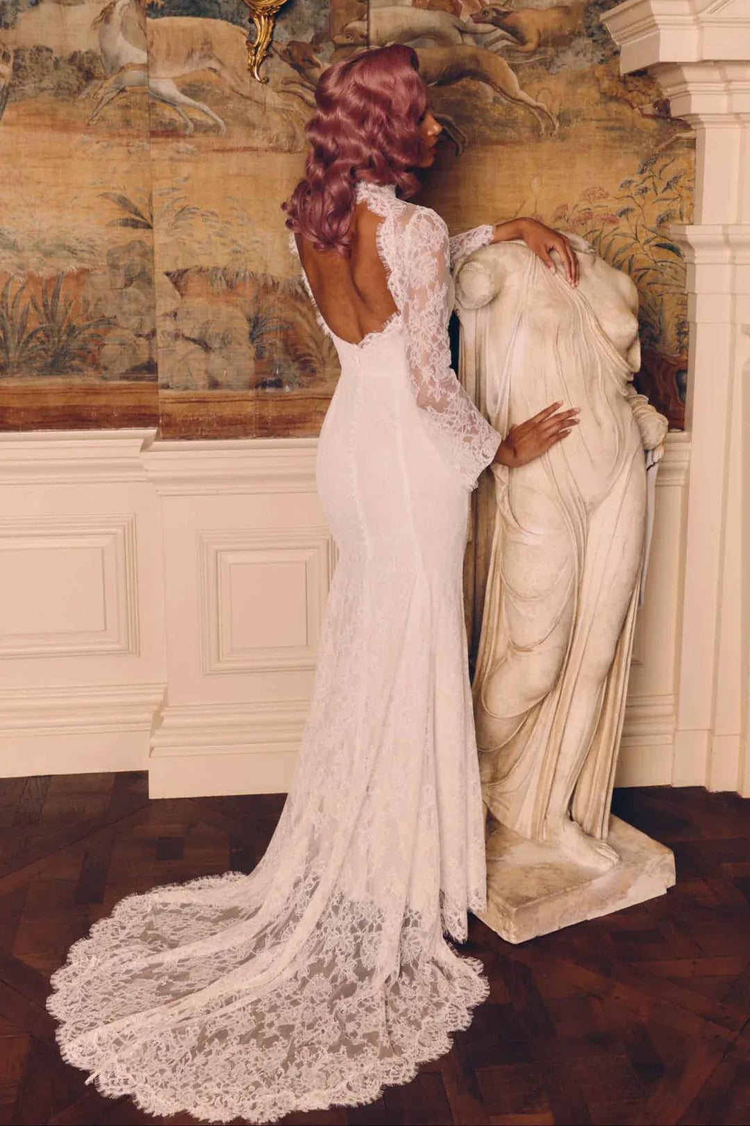 A woman wearing a fitted white lace wedding gown with long sleeves and an open back, posed indoors beside a classical statue in an ornate room with a large mural wall and dark wood floor.