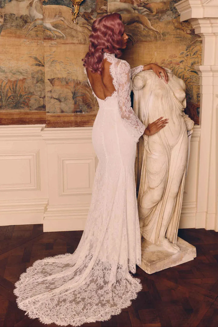 A woman wearing a fitted white lace wedding gown with long sleeves and an open back, posed indoors beside a classical statue in an ornate room with a large mural wall and dark wood floor.