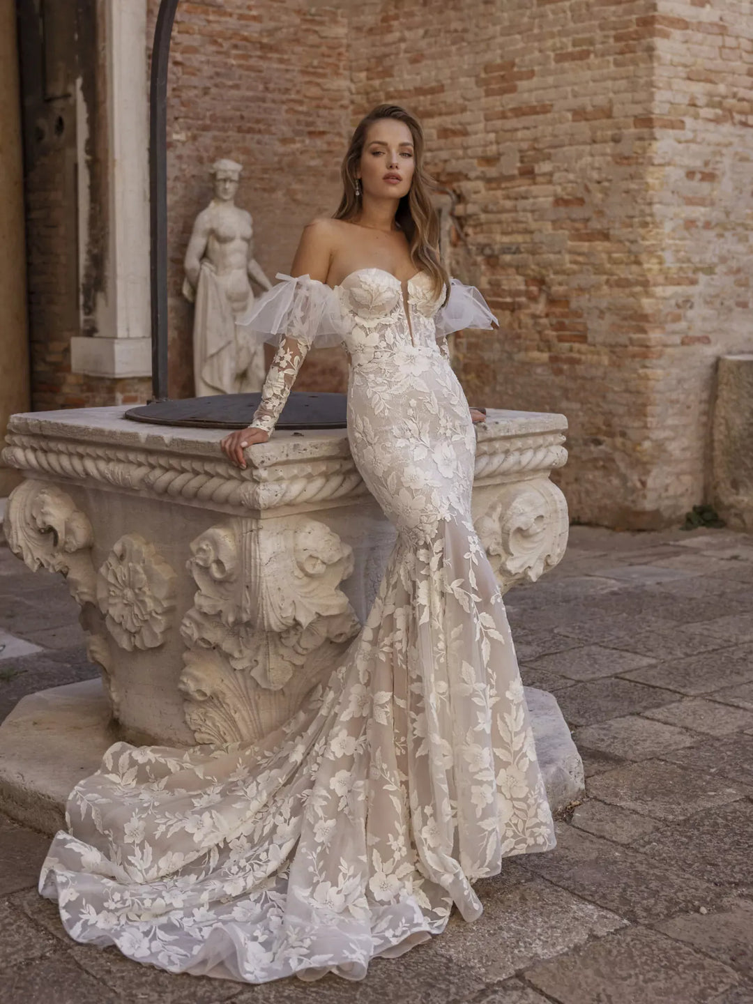 Woman in a white lace wedding dress standing in front of a stone fountain.