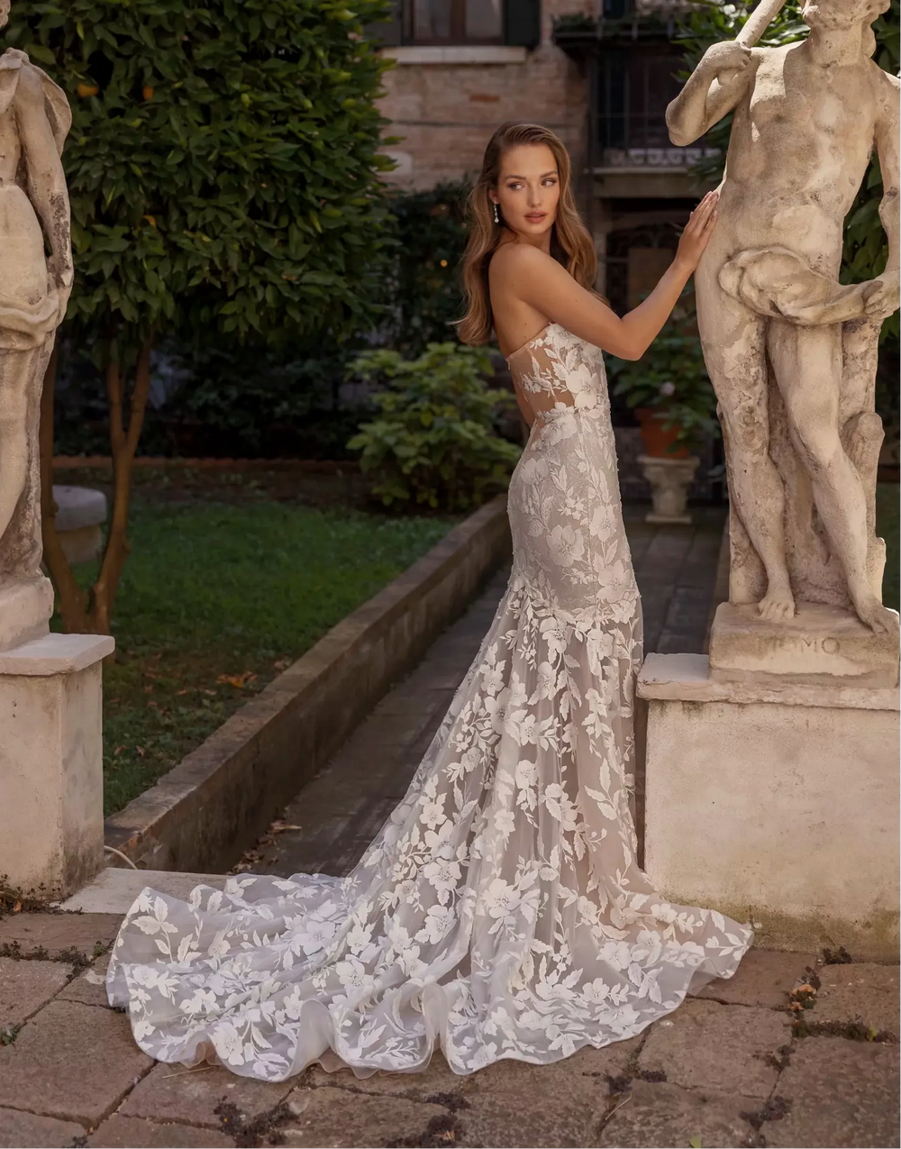 Woman in a lace dress standing between two classical statues in an outdoor setting.