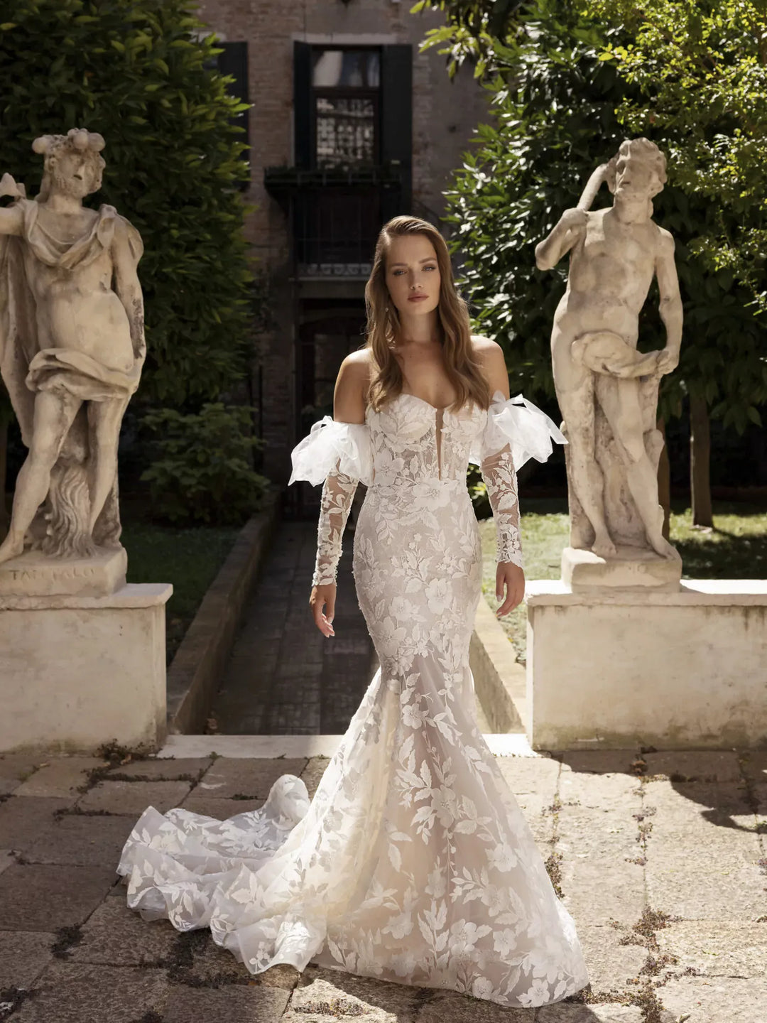 Woman in a white lace wedding dress standing between classical statues in an outdoor setting.