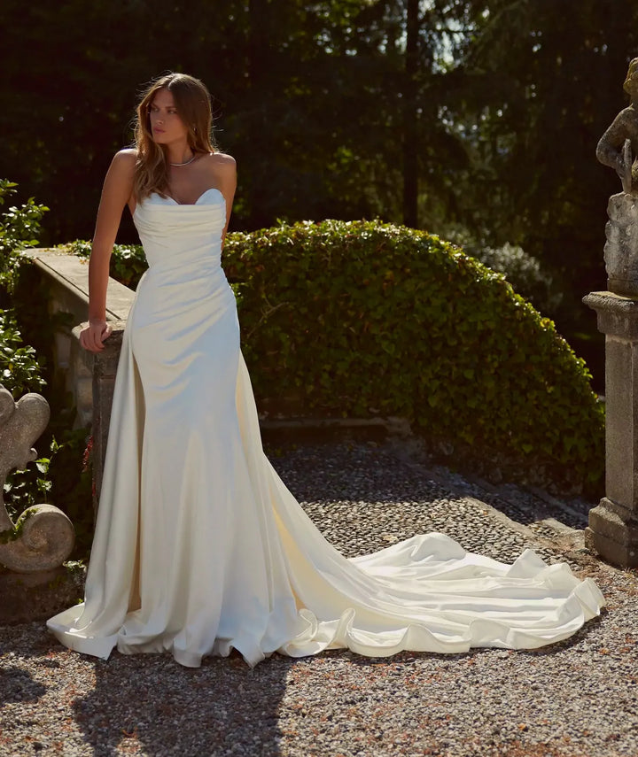 A woman wearing a strapless white wedding gown with a fitted bodice, draped skirt, and long train stands outdoors on a gravel path surrounded by greenery and stone statues.