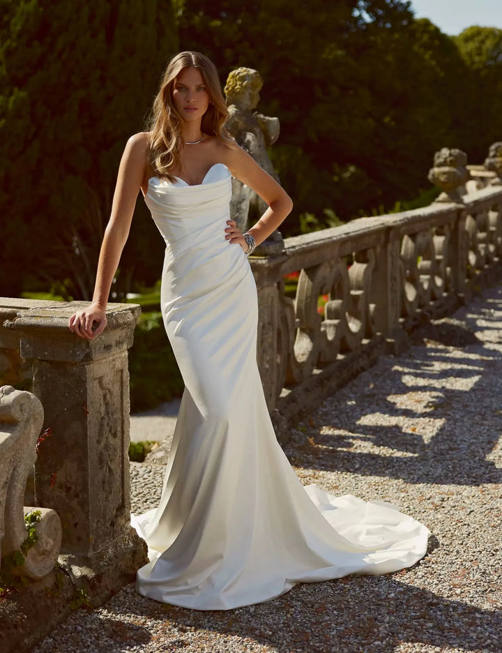 A woman wearing a strapless white satin mermaid-style wedding gown stands on a stone terrace outdoors, with trees and a statue in the background.