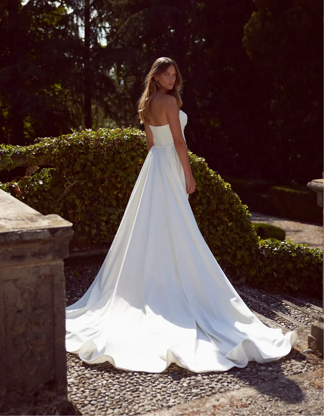 A woman wearing a strapless white wedding gown stands outdoors on a stone path with trimmed hedges and dark trees in the background. The dress has a fitted bodice and a full flowing skirt with a long train.