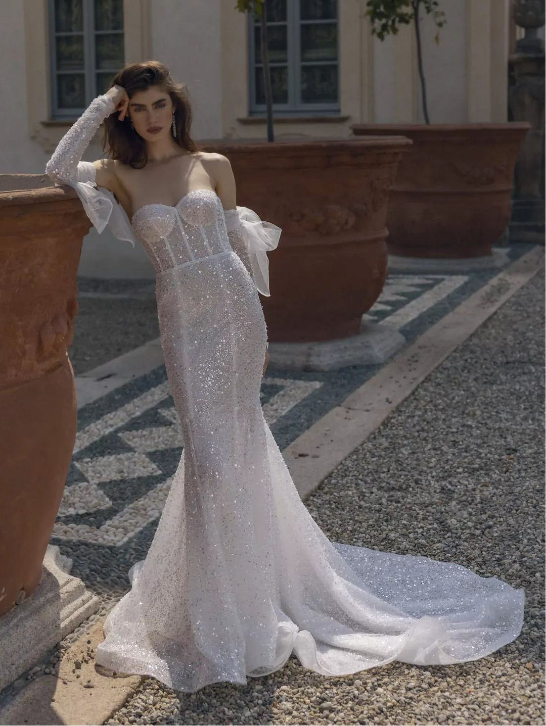 A woman models a sparkling white strapless mermaid-style wedding gown with sheer long sleeves, standing outdoors beside large terracotta planters in front of a building with windows.
